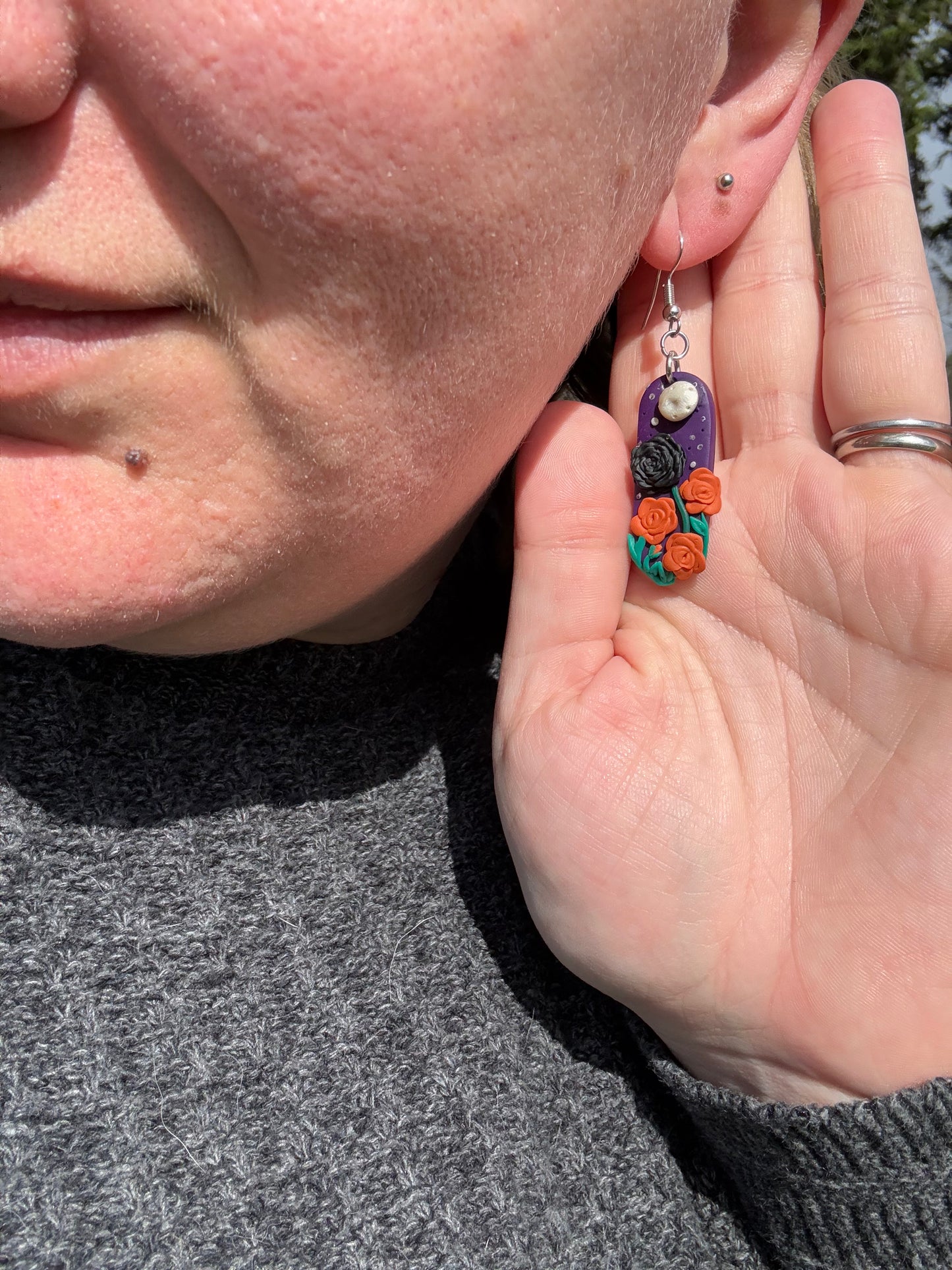 Capsule-shaped purple earrings with a full moon ornament, one black rose flower, and three orange rose flowers.