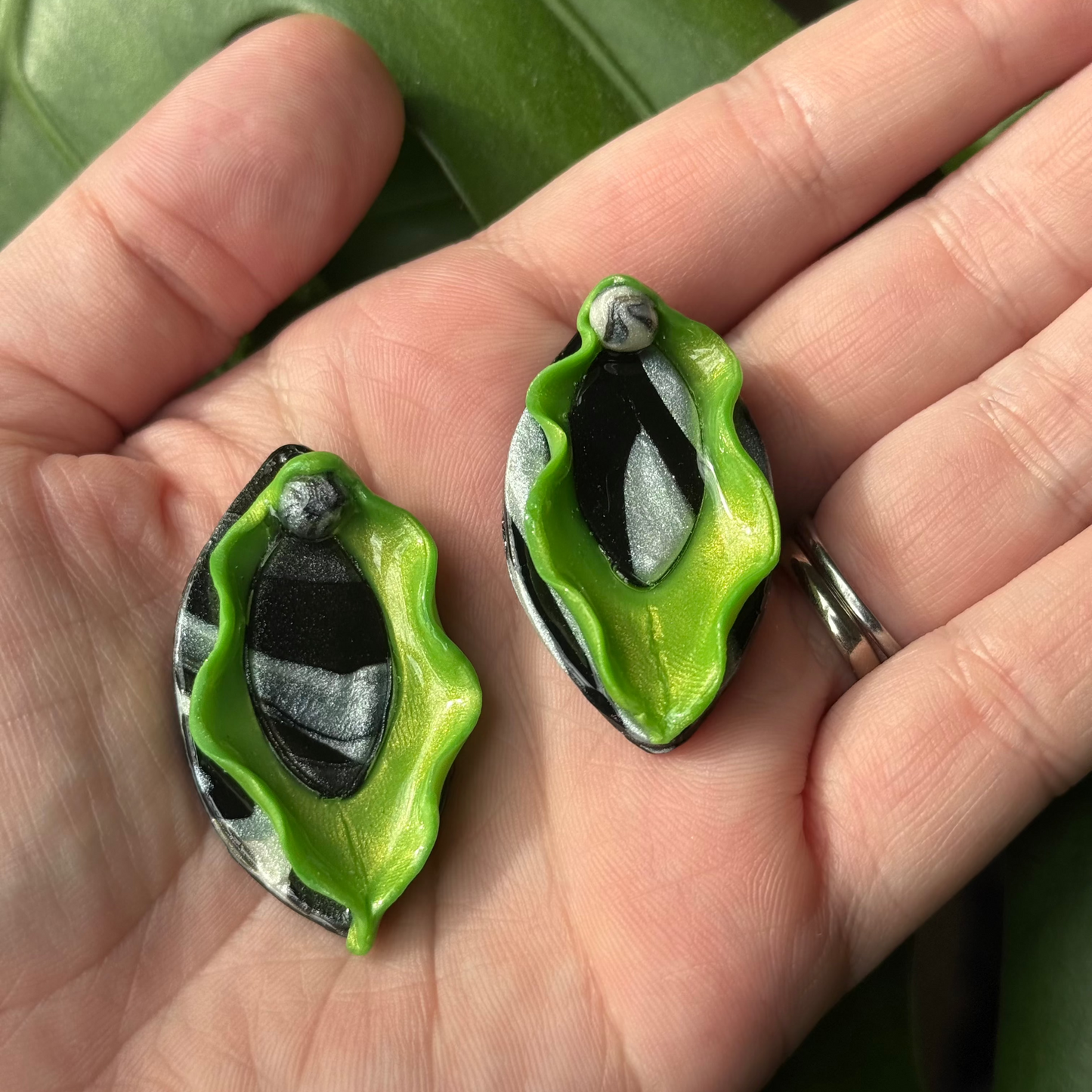 Green vulva dangle earrings with green labia minora and black clitoris and labia majora displayed in the palm of somebody's hand. Made of polymer clay.