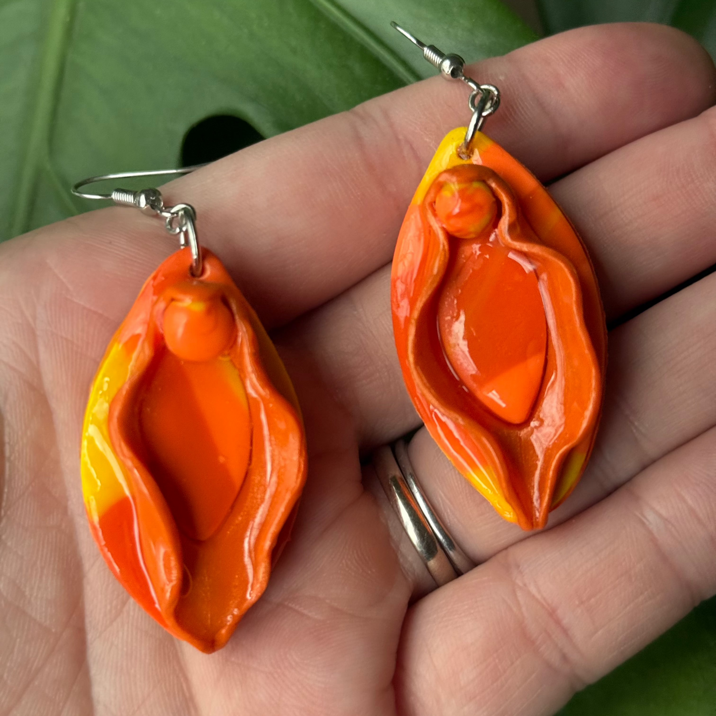 Orange and yellow marbled vulva dangle earrings are displayed in the palm of somebody's hand. Made of polymer clay.