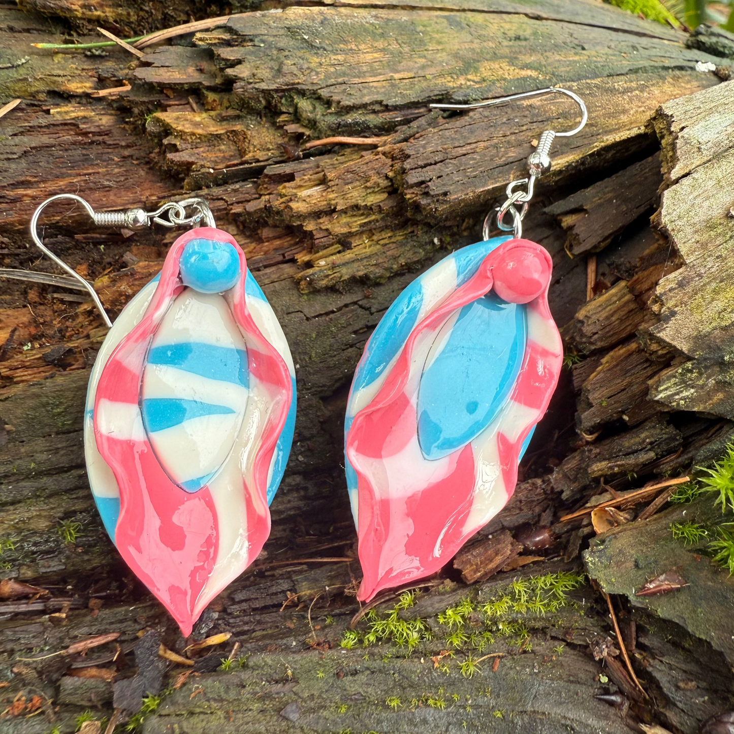 Marbled light pink and blue vulva dangle earrings resting against a tree trunk. Made of polymer clay.