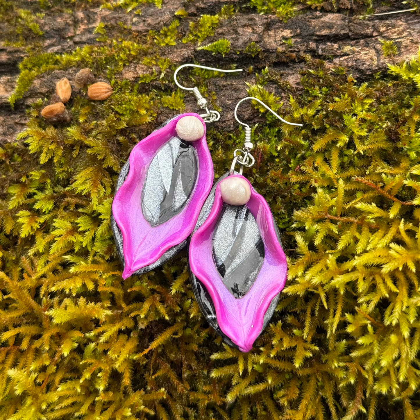 Purple vulva dangle earrings with purple labia minora and black clitoris and labia majora and silver hardware displayed on a mossy tree log. Made of polymer clay.