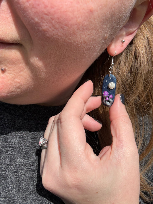 Capsule shaped black earring with a full moon ornament and three mushrooms with purple caps and white dots. The earring sparkles resembling a night sky.