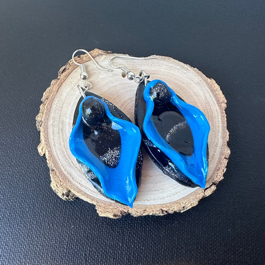 Blue vulva dangle earrings with blue labia minora and black labia majora and clitoris sitting on a tree log display.