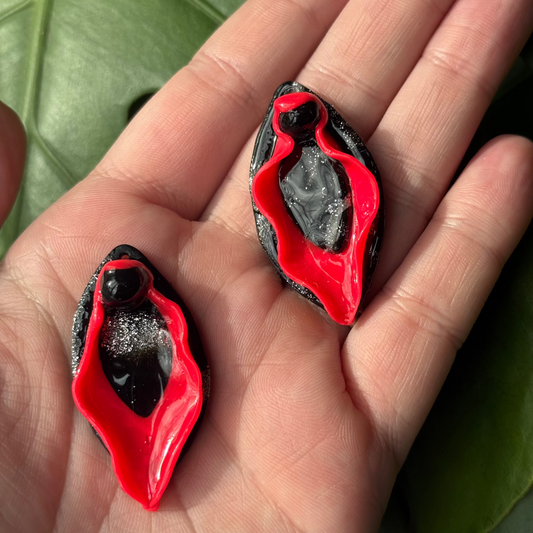 Red vulva dangle earrings with red labia minora and black clitoris and labia majora displayed in the palm of somebody's hand. Made of polymer clay.